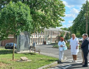 A kép bal oldalán az ápoló elődök emlékére ültetett kőrisfa látható, alatta az emléktáblával, háttérben az ápolók padjával. Jobb szélen a püspök és a delegáció néhány tagja áll.