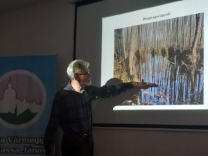 Tóth István Zsolt az előadás közben, az egyik diára mutatva beszél, a projektorról rávetülő megvilágítás ban