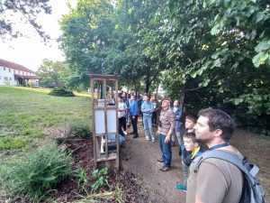 A program résztvevői a kórházpark egyik sétányán, a fák alatt állnak és felfelé tekintve hallgatják a madárhangokat és figyelik a madarak röptét.