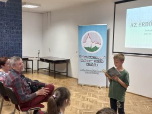 Az előadóteremben egy kisfiú könyvből olvas fel. a közönség hallgatja